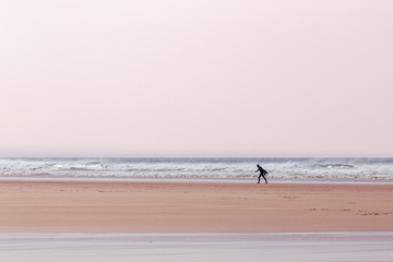 Fototapeta na wymiar Lone surfer, carrying a surfboard, on a beach in Cornwall, UK