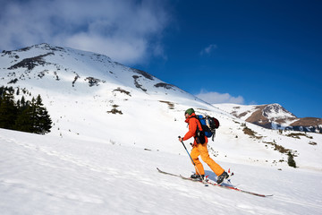 Skier tourist backpacker walking on skis up steep hill on background of bright blue sky and beautiful mountains ridges. Winter vacations, active lifestyle, skiing and trekking concept.