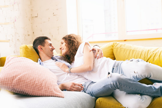 Young Happy Couple Lying And Hugging On The Yellow Couch In The Apartment, Love And Affection, A Young Family