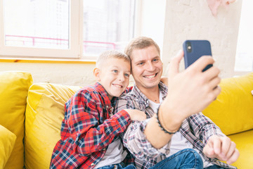 Young father with a son, a boy of 8 years old in checkered shirts, sitting on a yellow sofa, taking a selfie on a mobile phone, time together, relationship, family, one parent concept