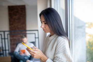Dark-haired wife standing near window using phone