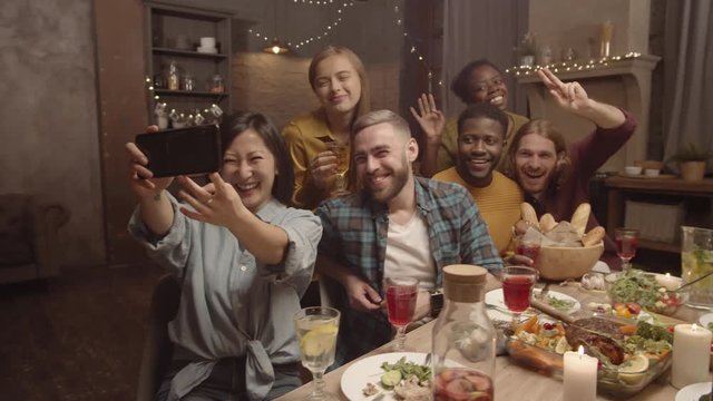 Handheld shot of happy Asian woman laughing and taking selfie with dinner party guests in her cozy home