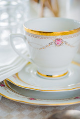 Vintage tea or coffee cup with gold rim on saucer. White linen tablecloth and napkin.
