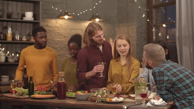 Handheld medium shot of happy group of friends toasting and clanking wine glasses while cooking food for dinner party in cozy kitchen