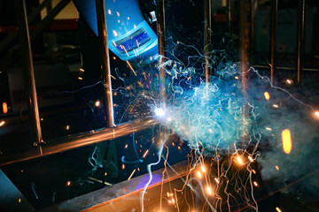 A welder at work in a workshop produces metal structures. Sparks from welding fly around.