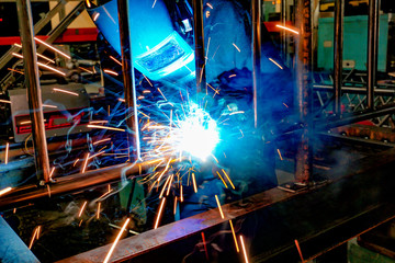 A welder at work in a workshop produces metal structures. Sparks from welding fly around.