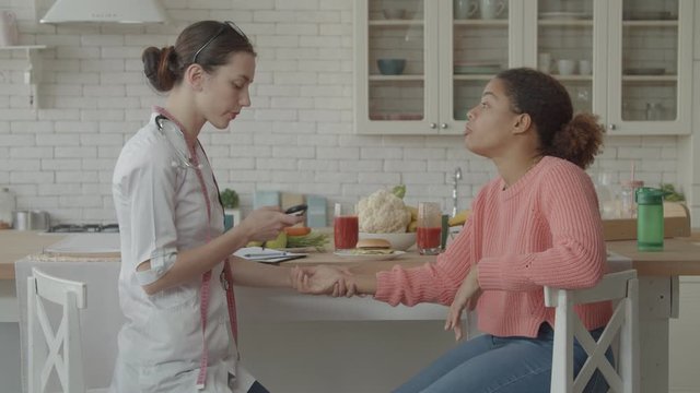 Beautiful caring female doctor checking pulse rate of worried patient in domestic kitchen. Empathetic physician telling bad news to african american woman after medical checkup at home.
