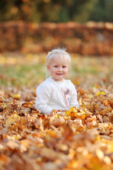 Little cute 3-4-year-old blonde girl sits on fallen leaves and smiles while walking in the autumn park