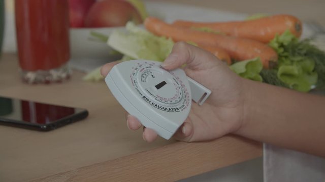 Close-up of female hand using body mass index or BMI calculator, measuring the relative weight based on an individual's mass and height at kitchen table with fresh healthy food in background.