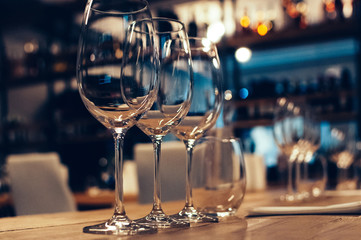 Empty glasses on wooden table, served for wine tasting event. Bar or shop interior, subdued light, lovely atmosphere, selective focus, film grain effect. Bottles on shelves in the background