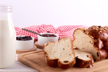 Still life with string of Christmas cake or sweet pastry with raisins and almond slices, glass of milk and bowls with fruit jam. Red kitchen towel on white background