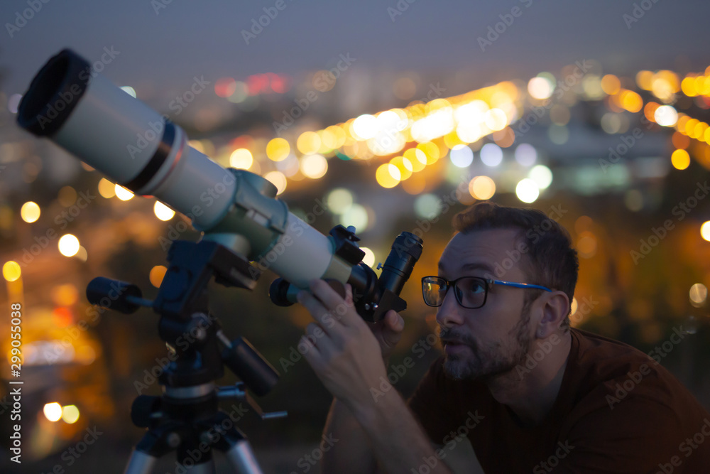 Wall mural astronomer with a telescope watching at the stars and moon with blurred city lights in the backgroun