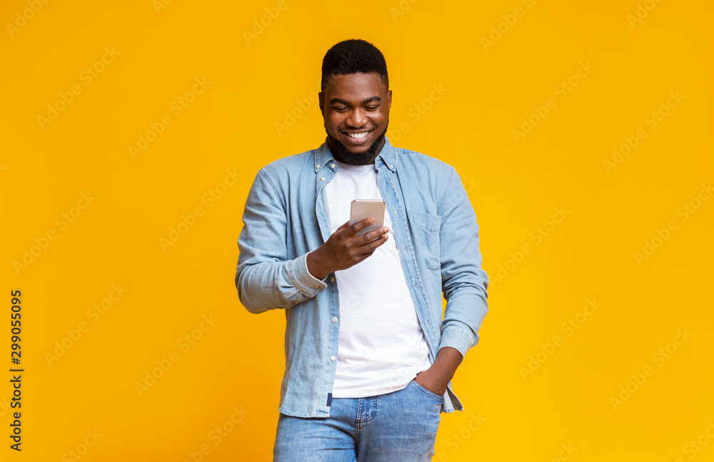 Poster cheerful black guy reading message on smartphone over yellow background