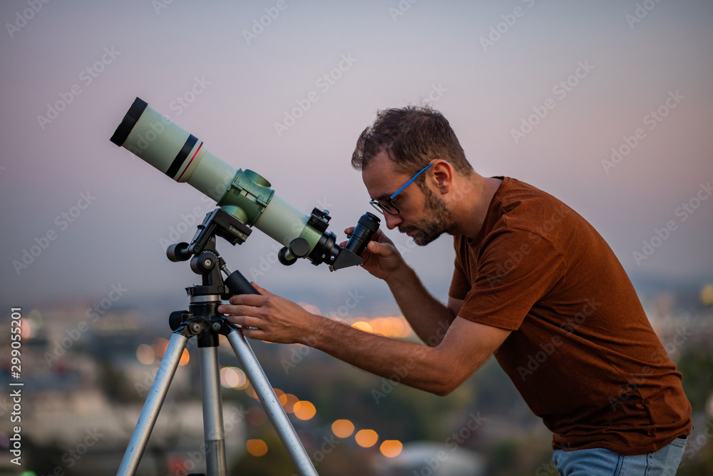 Wall mural astronomer with a telescope watching at the stars and moon with blurred city lights in the backgroun