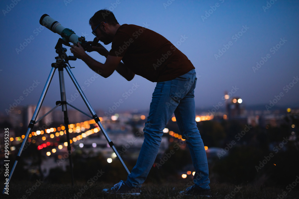 Wall mural astronomer with a telescope watching at the stars and moon with blurred city lights in the backgroun