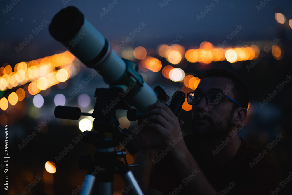 Wall mural astronomer with a telescope watching at the stars and moon with blurred city lights in the backgroun