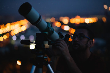 Astronomer with a telescope watching at the stars and Moon with blurred city lights in the...