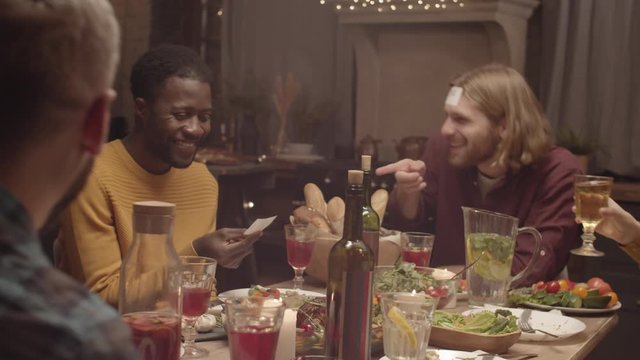 Handheld shot of happy people playing &laquo;Who am I&raquo; game at dinner party. They are laughing at African-American man looking at what is written on his sticky note, then clanking glasses