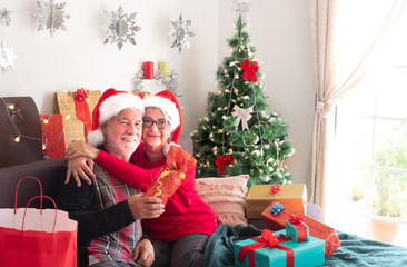 Obraz na płótnie Canvas Time of Christmas gifts. A senior couple of people smile happy looking at camera. Wearing Santa's hat. Christmas tree on the background. White wall
