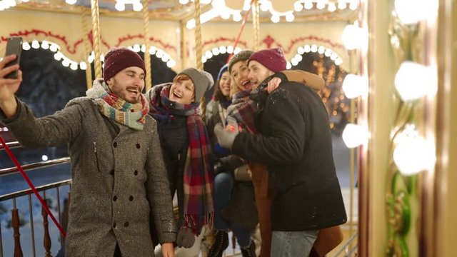 Happy people taking selfie photo on smartphone camera at New Year winter fair amusement park. Smiling friends have fun riding carousel at Christmas market. Blogger works. Garland lights on backdrop.