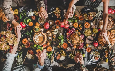 Fotobehang Eten Familie bidden hand in hand aan Thanksgiving tafel. Plat leggen van feestende mensen overhandigt Friendsgiving-tafel met herfstvoedsel, kaarsen, geroosterde kalkoen en pompoentaart over houten tafel, bovenaanzicht