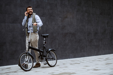 Fototapeta na wymiar Full length of cheerful handsome caucasian fashionable man listening music and pushing bicycle. In background is gray wall.