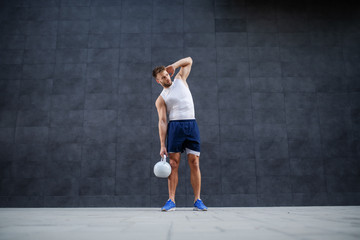 Full length of handsome strong muscular bearded Caucasian man in shorts and t-shirt posing with kettle bell while standing outdoors in front of gray wall.