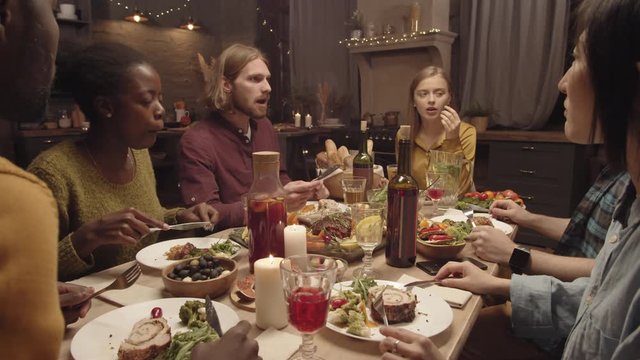 Handheld shot of multi-ethnic group of people eating and chatting while having dinner party in cozy kitchen