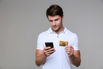 Concentrated pleased man holding debit card using mobile phone.