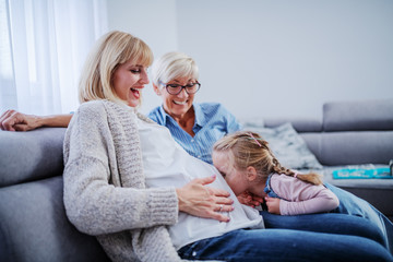 Smiling beautiful pregnant woman holding belly and sitting on sofa in living room while her daughter kissing belly. Next to pregnant woman is her cheerful proud mother.