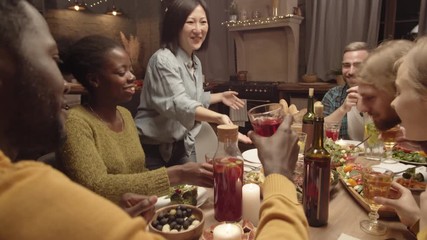 Handheld shot of happy Asian woman smiling and putting dish of roasted steak with vegetables on dinner table, then clanking glasses with group of cheerful friends and celebrating