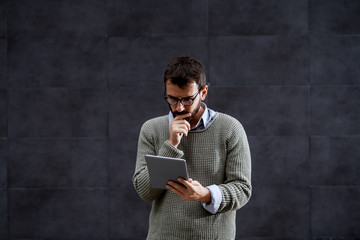Serious handsome caucasian stylish bearded man in sweater and with eyeglasses looking at tablet while standing outdoors in front of gray wall.