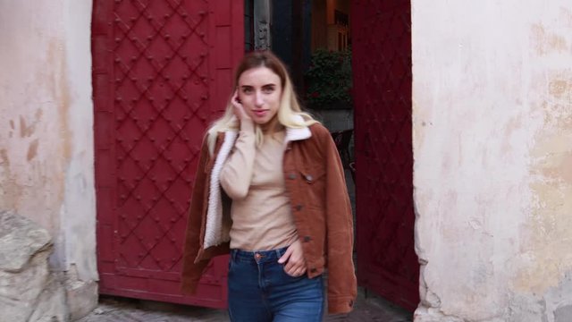 Stylish autumn fashionable video of a girl. Brown jacket with fur, jeans, boots, basic beige golf. Blonde on old city background