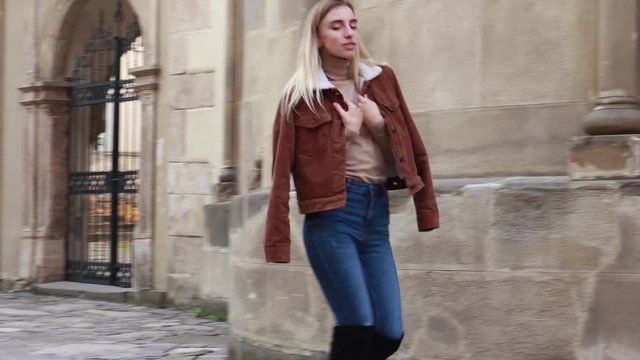 Stylish autumn fashionable video of a girl. Brown jacket with fur, jeans, boots, basic beige golf. Blonde on old city background