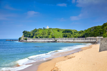 Fototapeta na wymiar Landscape with a sandy beach and embankment of Saint-Jean-de-Luz, green hill with white chapel on top (Basque Country, Atlantic coast, France). Coastal french town at sunny summer day. Sea shore