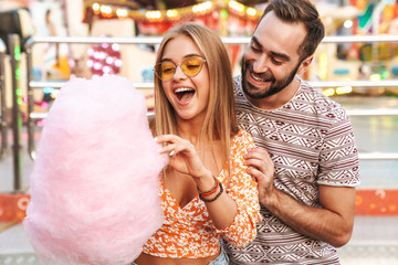 Positive loving couple in amusement park eat candyfloss.