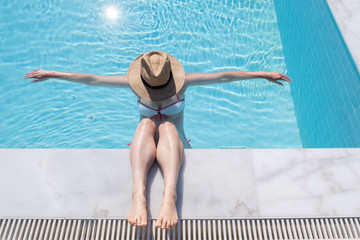 woman lying back in a swimming pool with calves on pool side wearing a hat