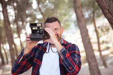 Hombre joven con camisa de cuadros haciendo fotos con una camara instantanea en el bosque.