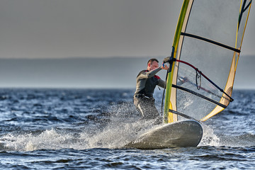 A male athlete is interested in windsurfing. He moves on a Sailboard on a large lake on an autumn...