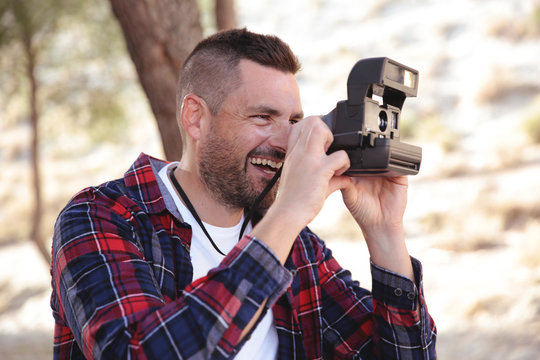 Hombre joven con camisa de cuadros haciendo fotos con una camara instantanea en el bosque.