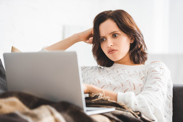 beautiful angry girl in blanket using laptop on sofa in cozy living room