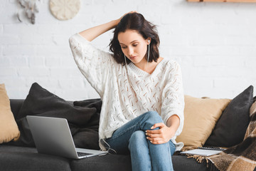 attractive girl thinking and studying online with notepad, earphones and laptop on sofa