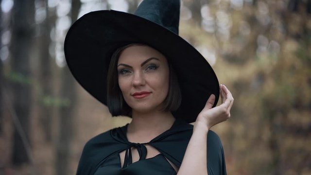 Portrait of young pretty witch in cap on autumn forest background. Halloween concept, cosplay dressing up. Slow motion