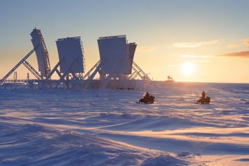 Fototapeta na wymiar Winter arctic landscape with large antennas of an abandoned troposphere communication station. Snowmobiles ride on a tundra covered with snow. Beautiful golden lighting at sunset. Chukotka, Russia.