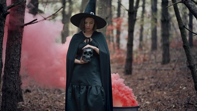 Black witch holding skull in hands, stroking it on autumn smoke forest background. Horror, halloween, cosplay holiday, magic concept