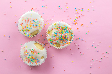 Three little white frosted white cupcakes in centre of pink background sprinkled with multi - coloured sprinkles on pink with copy space - top view
