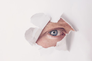 An eye of gray-blue color looks through a hole in white paper. The concept of secrecy, peeping....