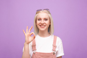 Happy teen girl shows a sign OK looks at the camera and smiles, wears a cute casual clothing and sunglasses, isolated on a purple background. Portrait of a happy teenager.