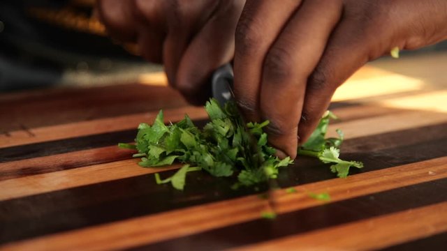 Close up slow motion shot of an African man&rsquo;s hands using a knife to chop up a bunch of parsley on a striped, wooden cutting board while wearing a white, shell bracelet on his wrist.
