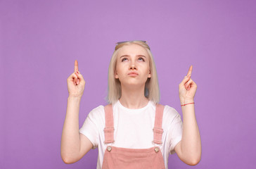 Funny girl in a pretty casual dress is standing against the background of a purple wall, looking up and showing her fingers on copyspace. Teen girl is suspicious looking up and showing her fingers
